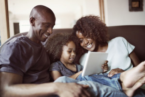 A photo of happy family sitting on sofa at home. Smiling parents are looking at son using digital tablet in living room. They are in casuals.