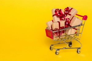 shopping cart with gifts on a yellow background. Gifts wrapped in kraft paper with a red ribbon and bow.