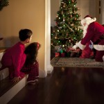 Two children peek at Santa Claus placing gifts under the Christmas tree.