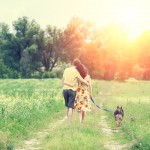 Happy couple in love with dog walking on a rural dirt road in springtime at sunset. The woman and man hugging. The woman keeps her dog on a leash. Couple and dog back to the camera.