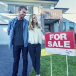 Young couple in front of a home with a For Sale sign.